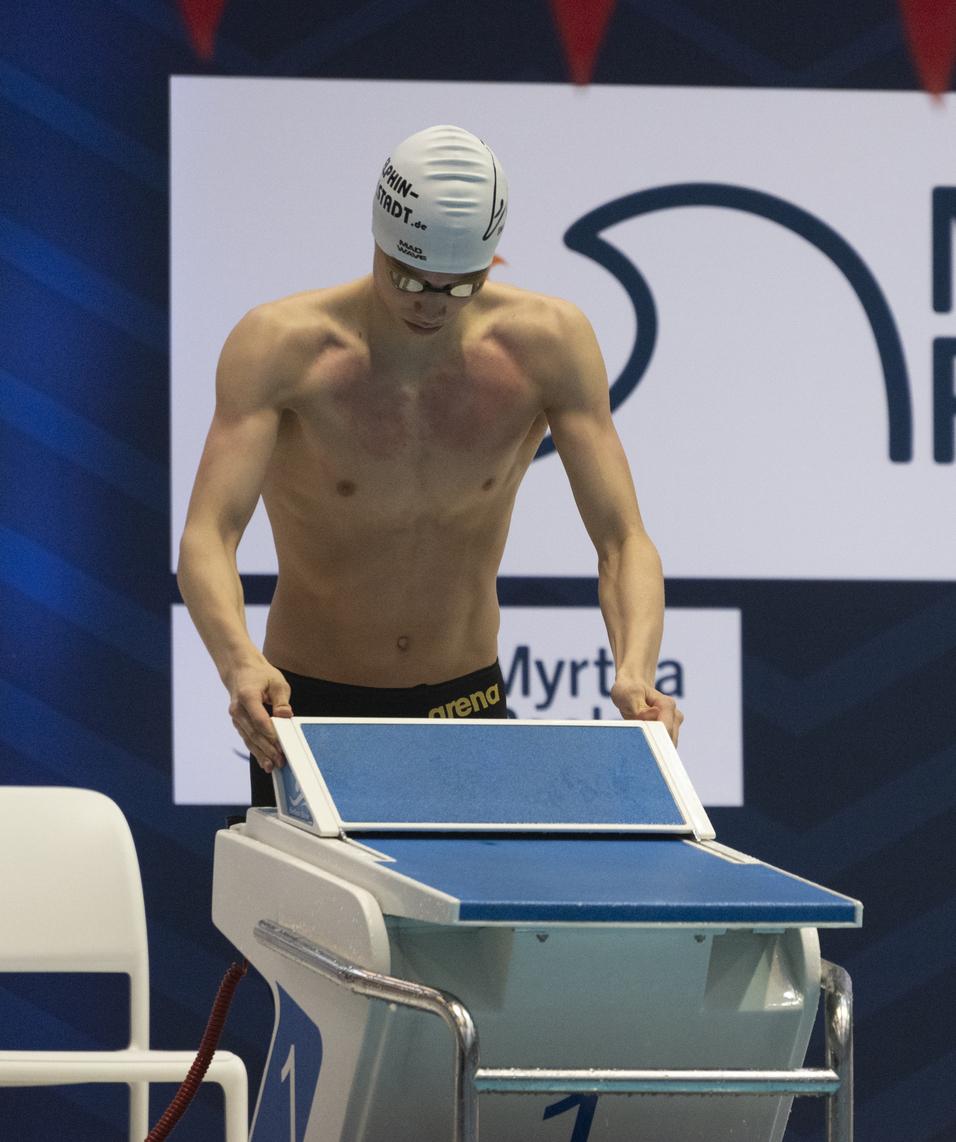 Schwimmer in Badehose mit Schwimmhaube beugt sich über einen Startblock vor einem Myrtha Pools Banner.