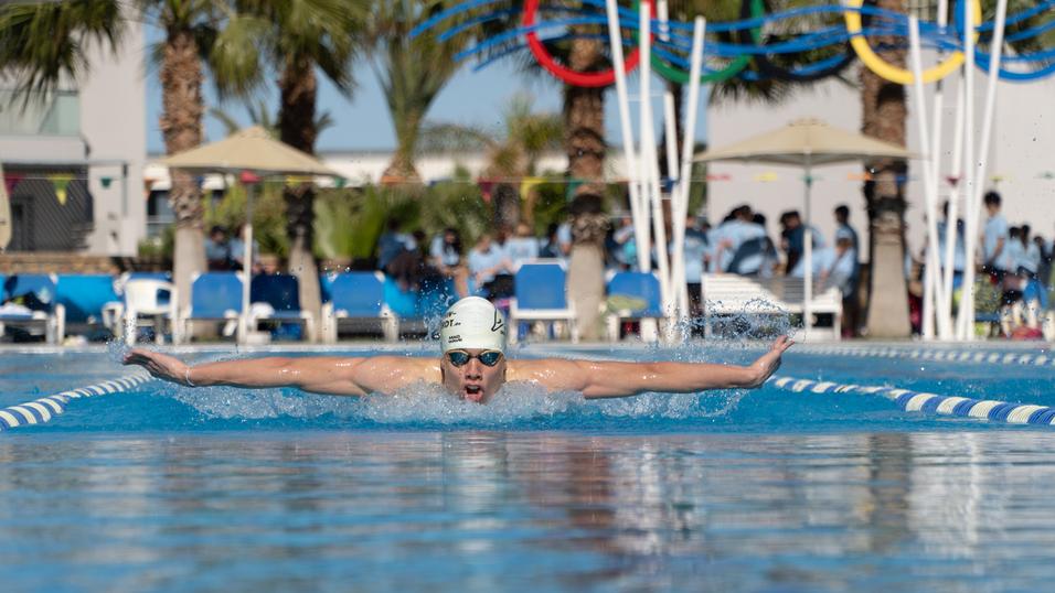 Schwimmer in einem weißen Badekappe zieht beim Schmetterlingsstil durch das Wasser einer blauen Schwimmhalle.
