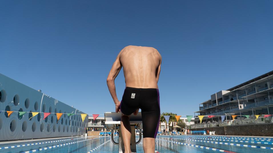 Sportler in Badehose steht am Startblock eines Schwimmbeckens unter klarem Himmel. Bunte Wimpel hängen über dem Wasser.
